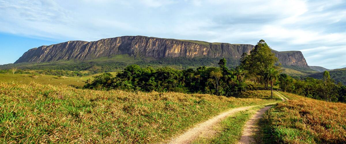 A dirt path leading to a large mountain with a flat top, surrounded by green vegetation and trees.