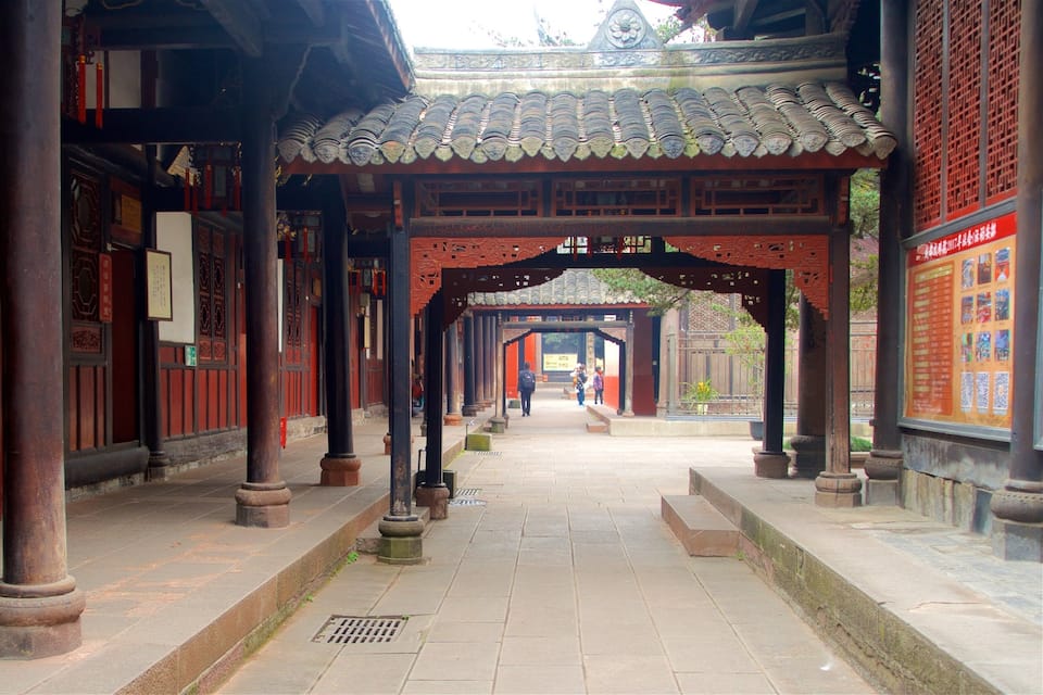 A traditional Chinese architectural corridor with red wooden doors and columns, leading to a tiled roofed pavilion.