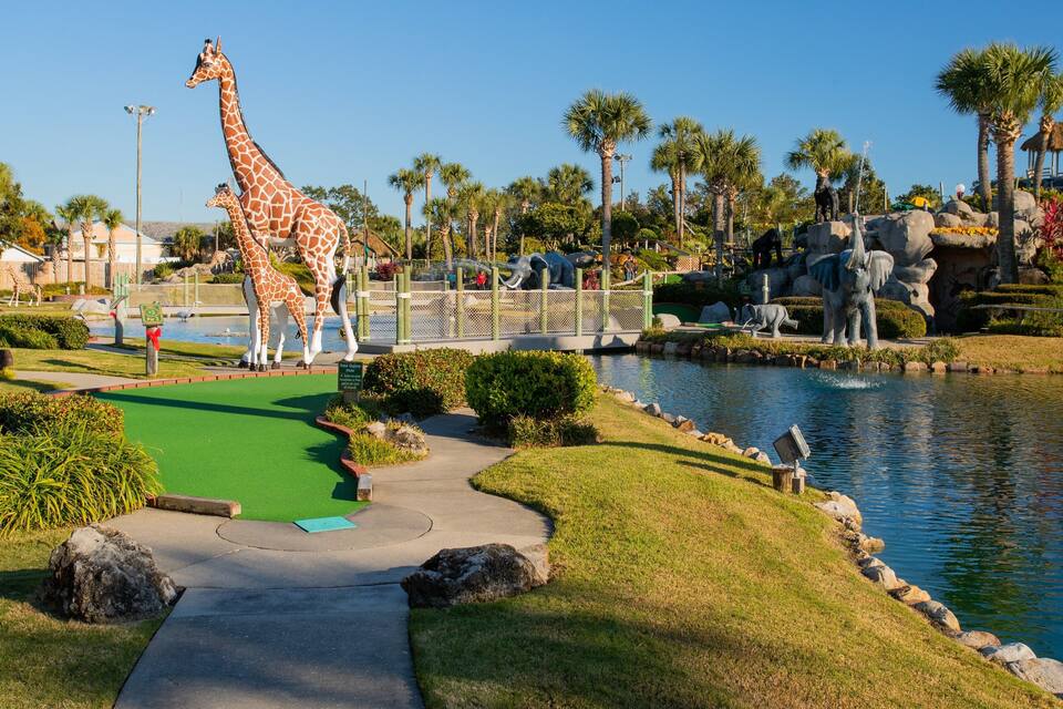 A park with a large giraffe statue, palm trees, a pond, and various animal sculptures.