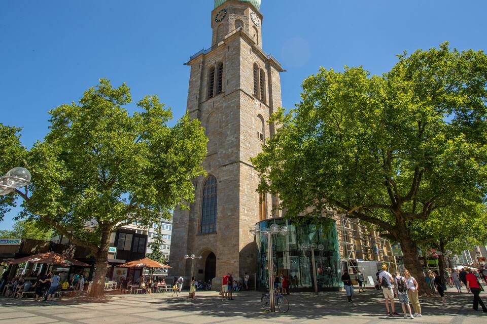 Une tour historique dotée d’une horloge, entourée d’arbres et bordée par une place publique où se promènent des gens.
