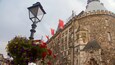A historic stone building with a clock tower, red flags, and a street lamp.