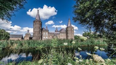 A historic building with multiple towers and domes, surrounded by lush greenery and a calm pond.