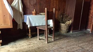 A rustic interior with wooden floors, a table with a white cloth, a chair, and a basket of dried plants.