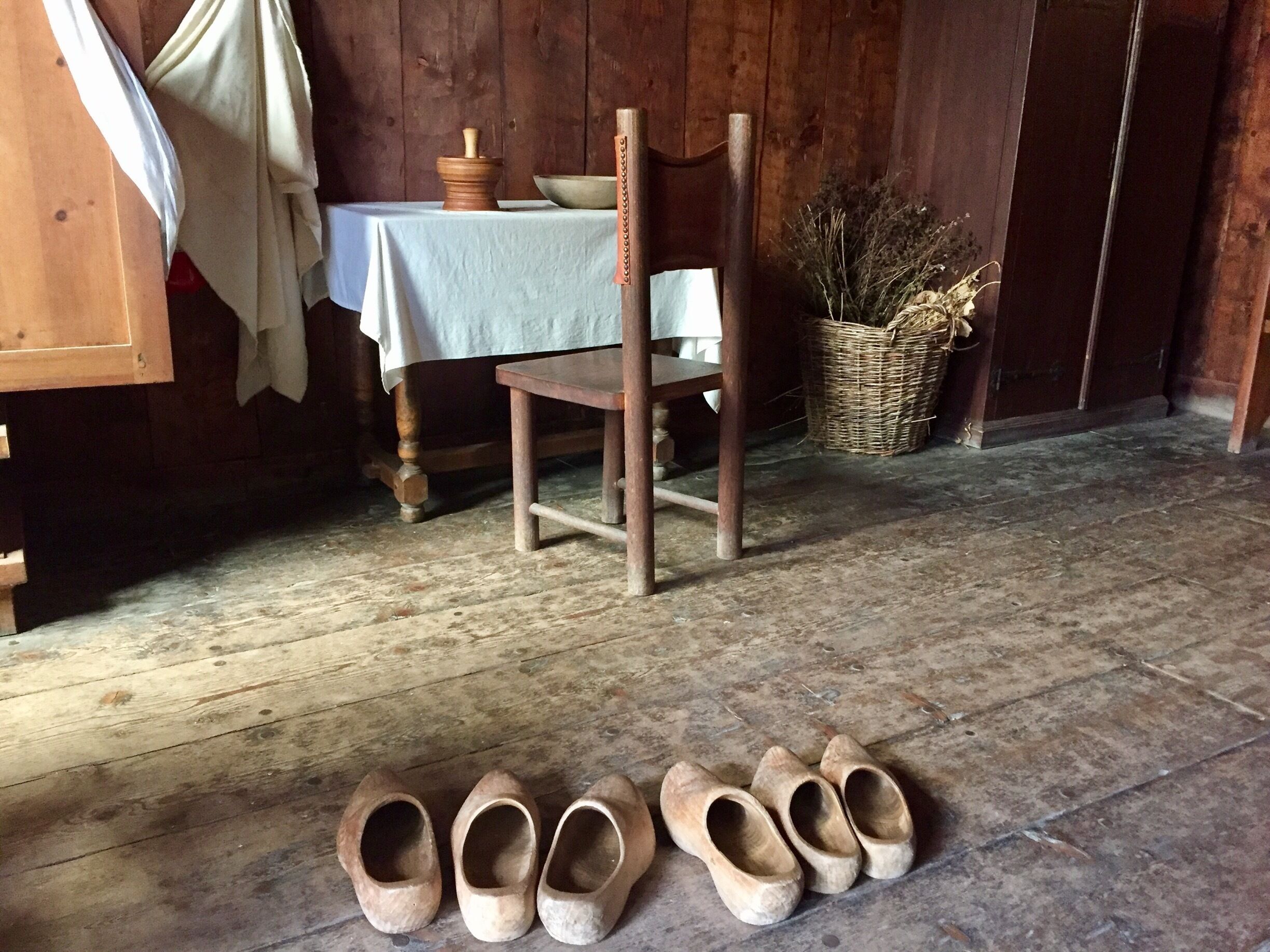 A rustic interior with wooden floors, a table with a white cloth, a chair, and a basket of dried plants.