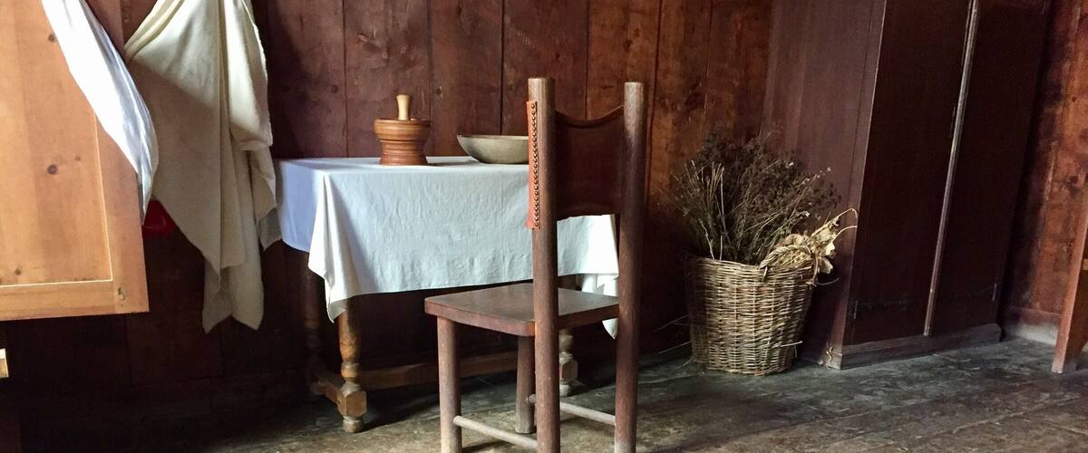A rustic interior with wooden floors, a table with a white cloth, a chair, and a basket of dried plants.
