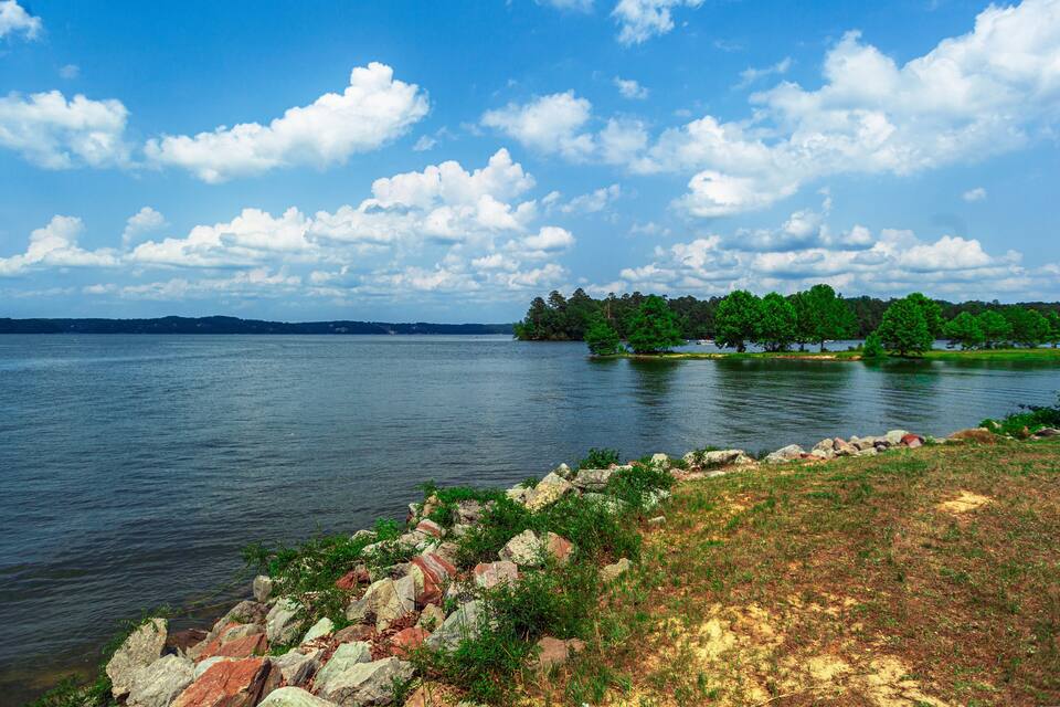 A calm lake with a rocky shoreline, a small island with trees, and a clear blue sky with clouds.