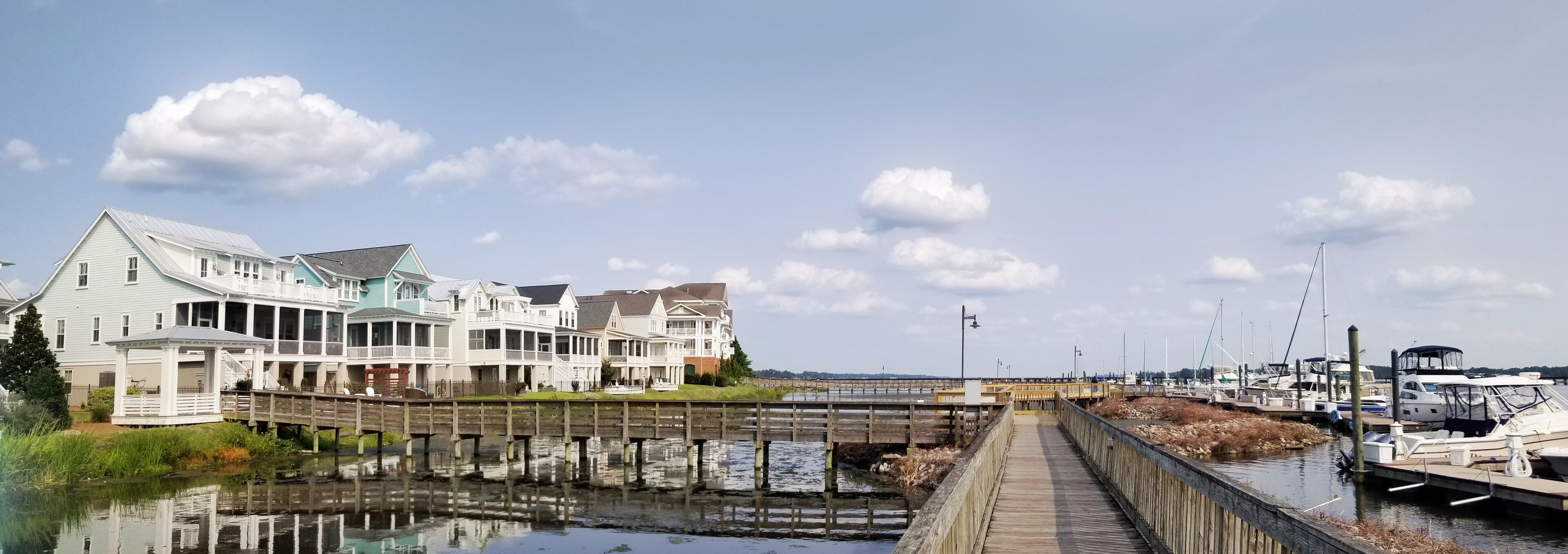 A waterfront with a wooden pier, a row of beach houses, and a marina with boats.