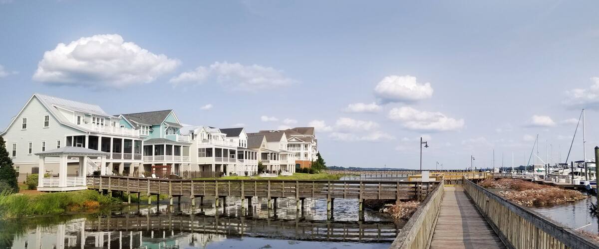 A waterfront with a wooden pier, a row of beach houses, and a marina with boats.