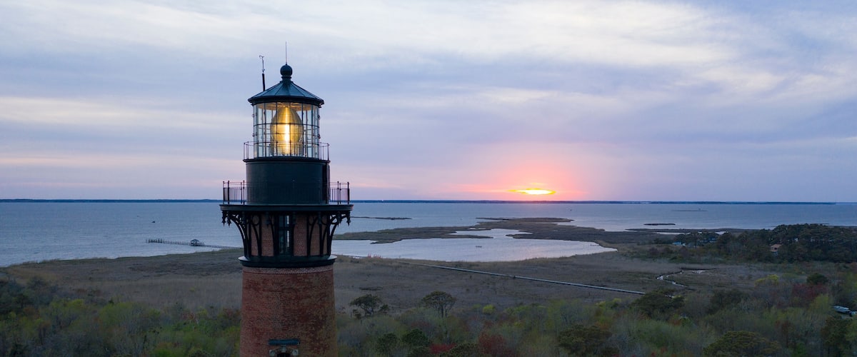 A lighthouse at dusk with the sun setting over a coastal landscape.