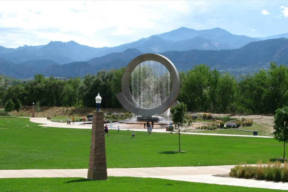 A circular water feature with a stone pedestal and a lamp post, surrounded by a grassy park with mountains in the background.