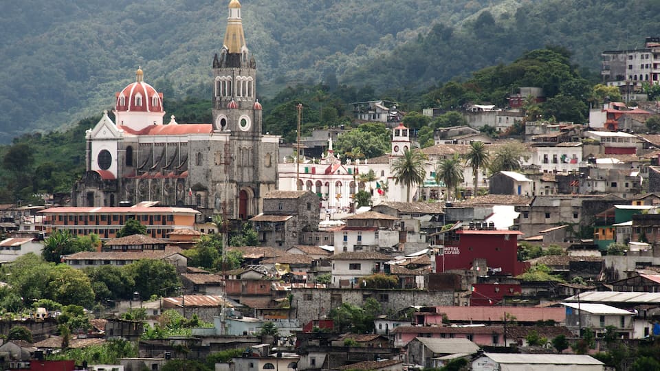 A historic church with a bell tower, surrounded by a densely populated hillside town with various buildings and lush greenery.