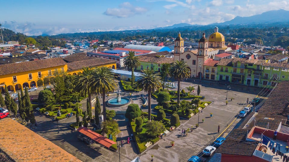 A cityscape with a prominent yellow building, a large yellow dome, and a fountain in a central square surrounded by palm trees and other greenery.