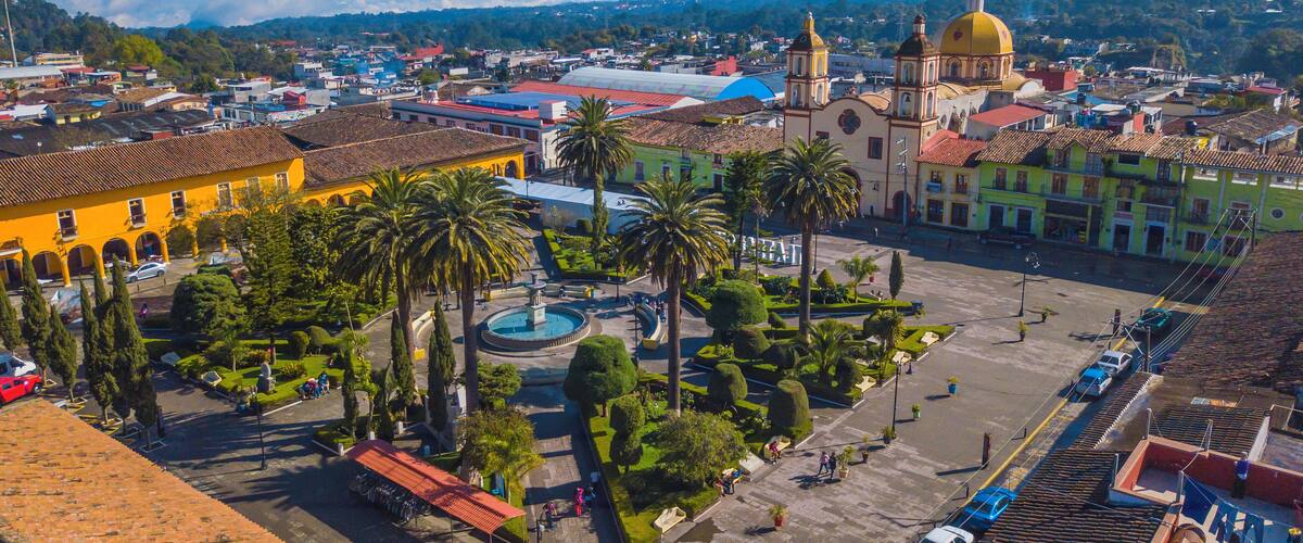 A cityscape with a prominent yellow building, a large yellow dome, and a fountain in a central square surrounded by palm trees and other greenery.