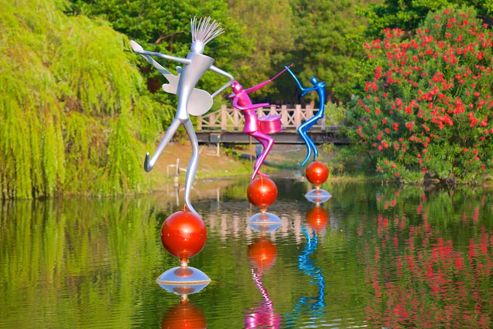 A sculpture of a figure balancing on a ball by a pond with red flowers and greenery in the background.