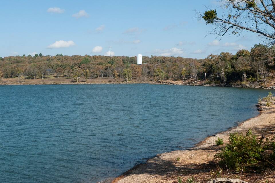 A calm lake with a sandy shore, surrounded by trees and a distant white building.