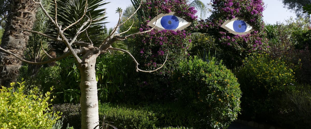 A garden with palm trees, purple flowers, and two large eye-shaped sculptures.