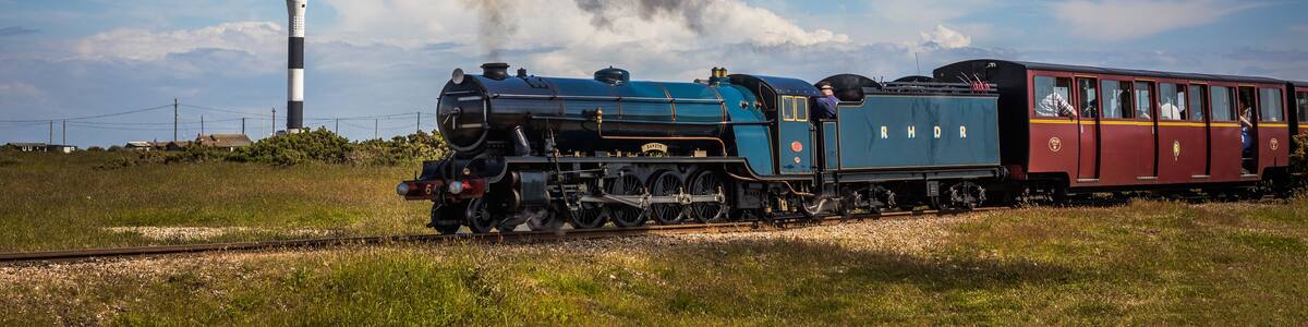 Eine Dampflokomotive mit Fahrgästen auf Gleisen, Rauch stiegend, mit einem Leuchtturm im Hintergrund.