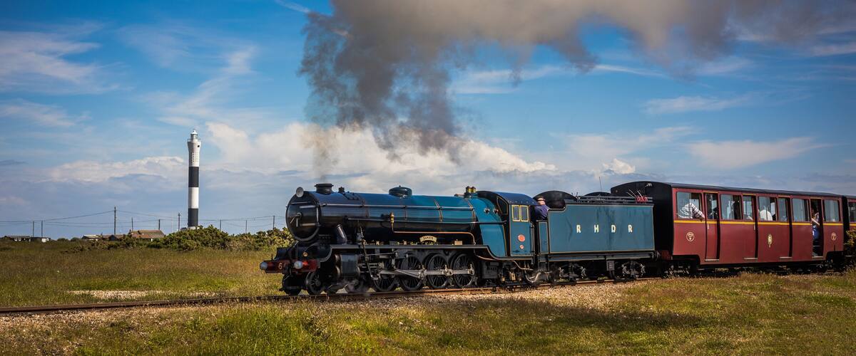 Una locomotora a vapor con pasajeros, expulsando humo, sobre vías de tren con un faro al fondo.