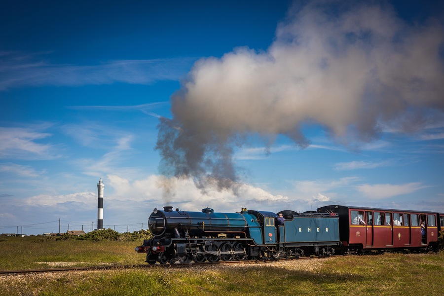 Una locomotora a vapor con pasajeros, expulsando humo, sobre vías de tren con un faro al fondo.