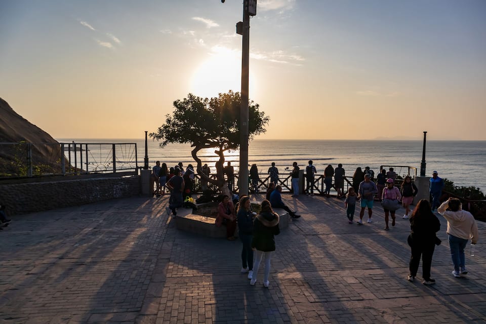 A coastal promenade with people walking and sitting near the water during sunset.