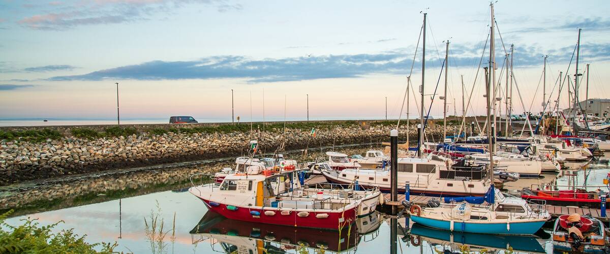 A marina with several boats docked, a rocky breakwater, and a van on the shore.