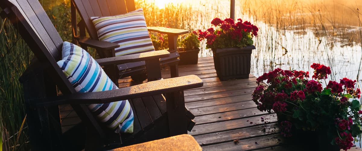 A wooden deck with two Adirondack chairs, a striped pillow, and a potted plant with red flowers.