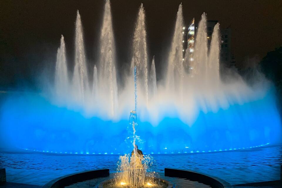 A nighttime fountain with water jets illuminated by blue lights, set against a cityscape.