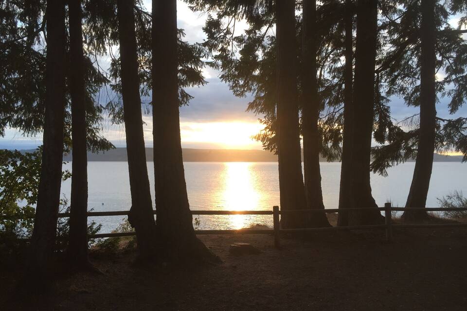 Um pôr do sol sobre um lago calmo, com árvores altas ao primeiro plano.