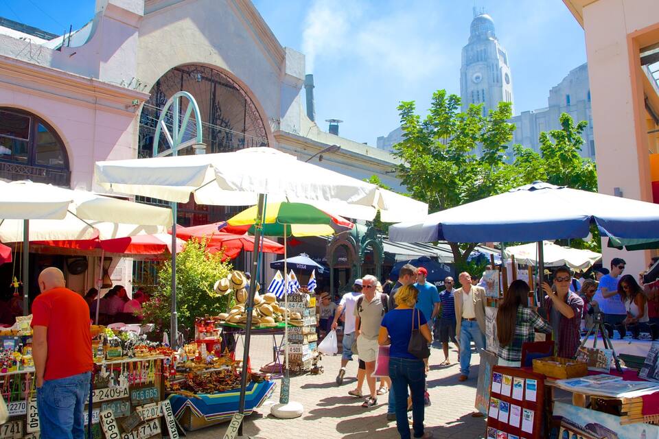 A bustling outdoor market with various stalls and umbrellas, surrounded by historic buildings and a prominent clock tower.