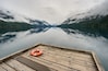 A wooden dock with a lifebuoy, surrounded by a calm lake reflecting mountains and forested areas.