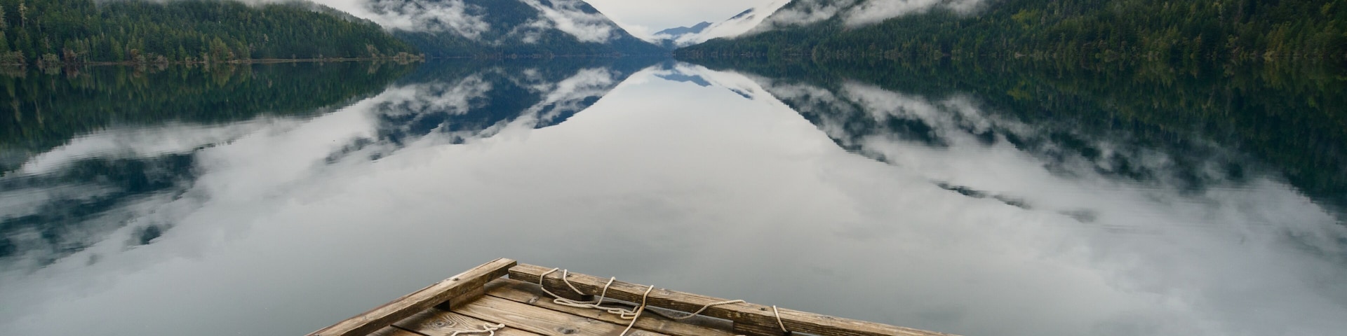 Eine hölzerne Anlegestelle mit einem Rettungsring, umgeben von einem stillen See, der Berge und bewaldete Gebiete spiegelt.
