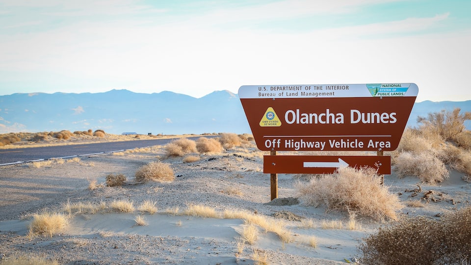 A sign for Olancha Dunes, an off highway vehicle area, with a desert landscape and mountains in the background.