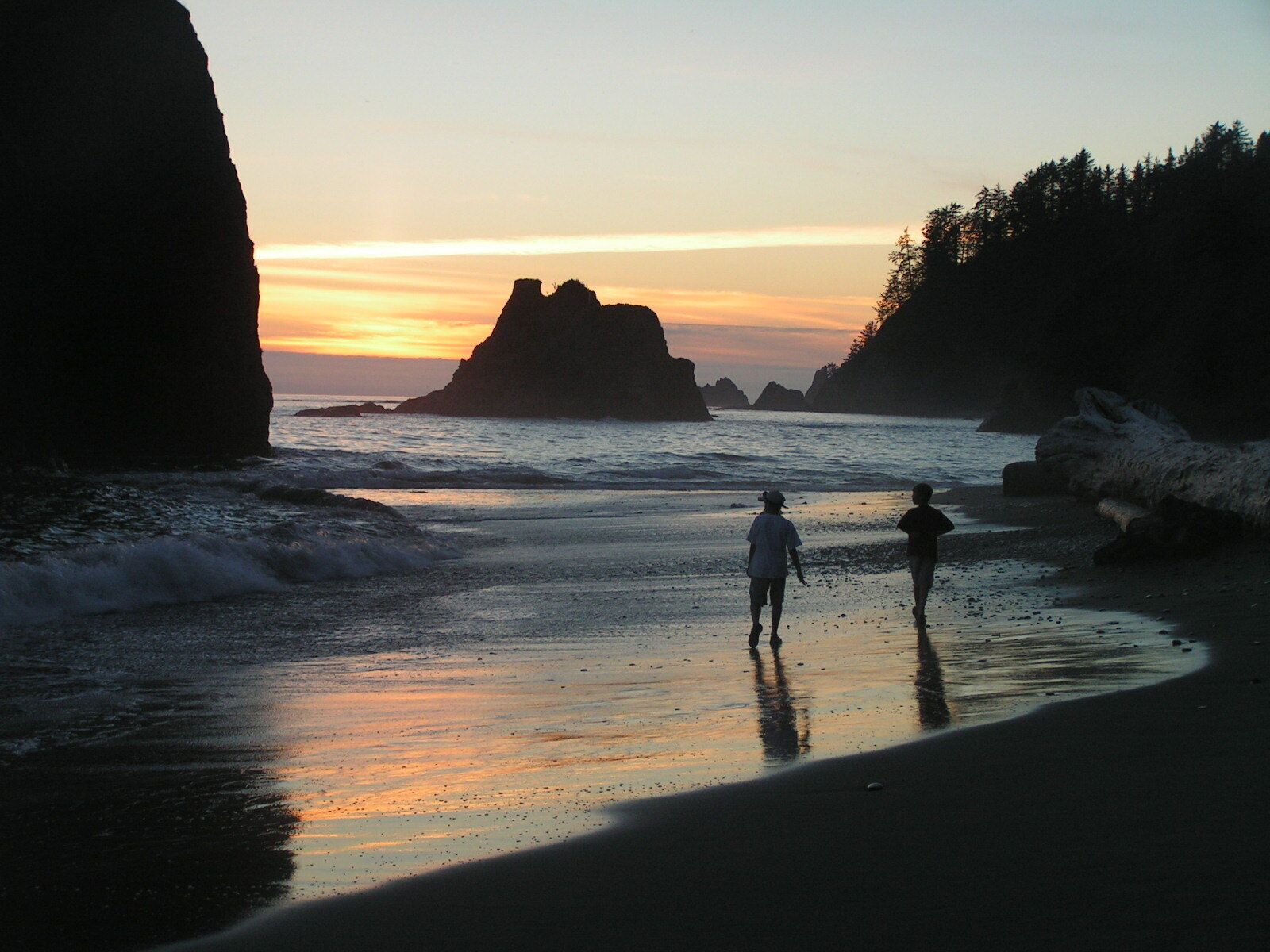 A beach at sunset with two people walking along the shore, surrounded by rocky cliffs and ocean waves.