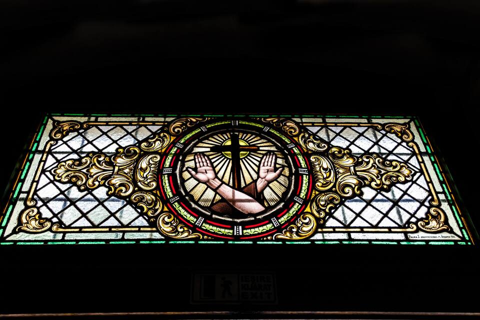 A stained glass window with a central circular design and hands reaching towards each other.