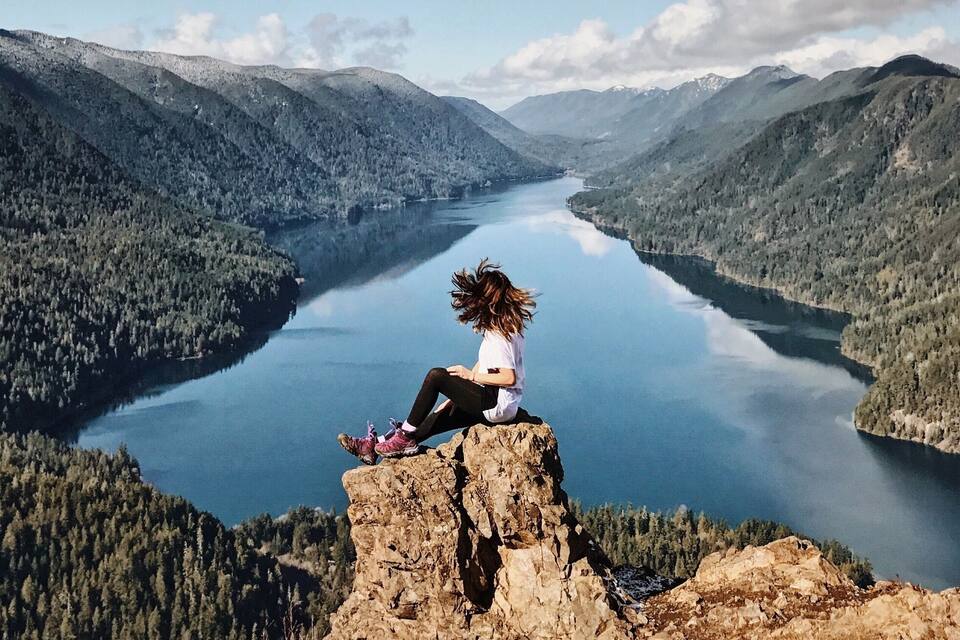 Uma pessoa sentada numa rocha com vista para um lago rodeado por montanhas florestadas.