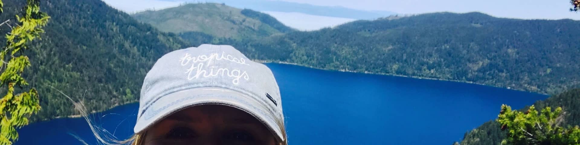 A person wearing a cap with the text "tropical things" taking a selfie with a scenic view of a lake surrounded by forested hills.