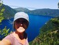 A person wearing a cap with the text "tropical things" taking a selfie with a scenic view of a lake surrounded by forested hills.