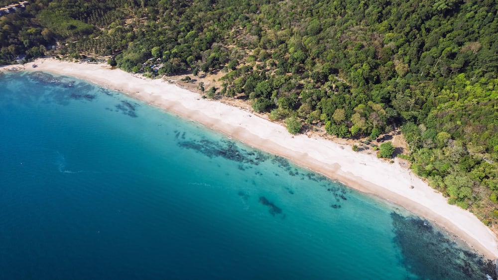 A coastal landscape with a sandy beach, clear turquoise waters, and dense green forest.