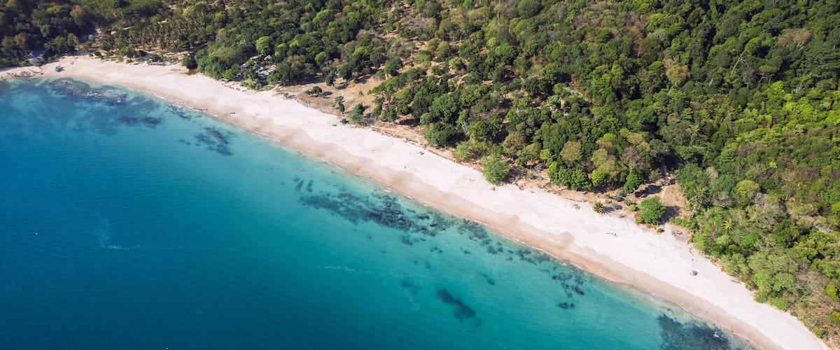 A coastal landscape with a sandy beach, clear turquoise waters, and dense green forest.