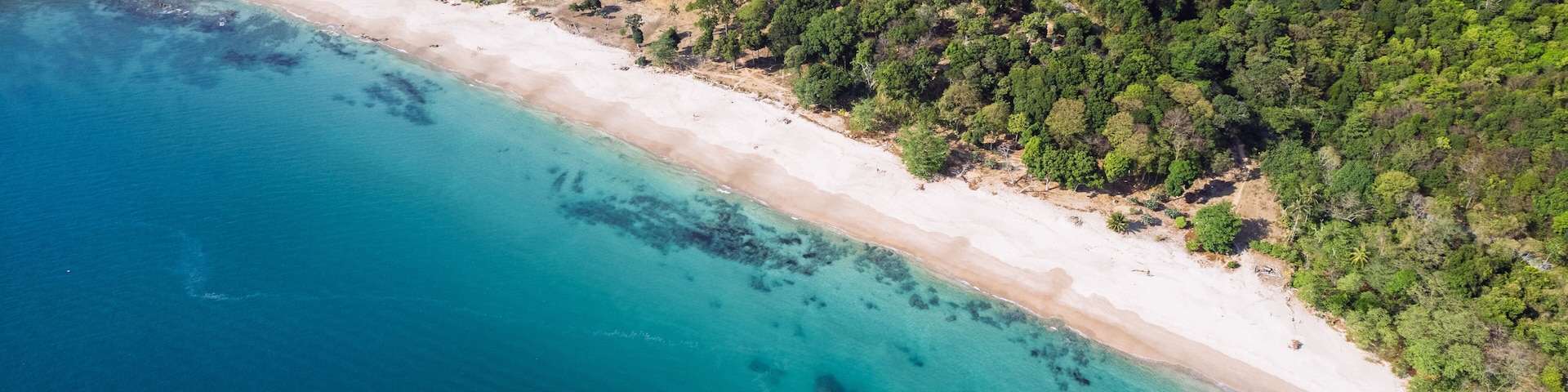 A coastal landscape with a sandy beach, clear turquoise waters, and dense green forest.