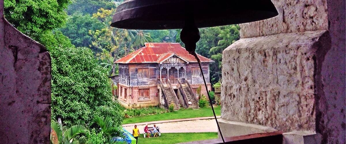 A large bell hanging from a structure, overlooking a building with a red roof surrounded by lush greenery.