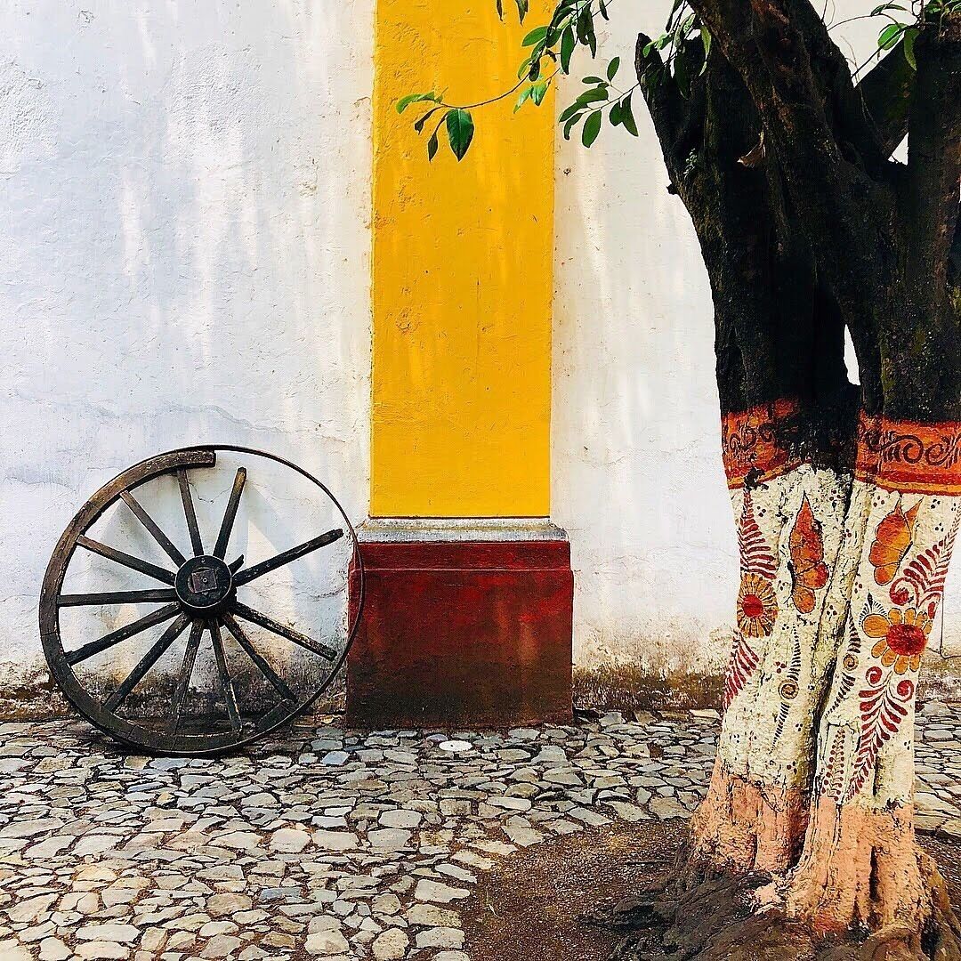 A cobblestone path, a yellow column, a wooden wheel, and a tree with patterned fabric.