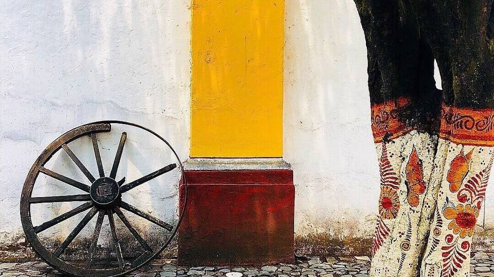 A cobblestone path, a yellow column, a wooden wheel, and a tree with patterned fabric.