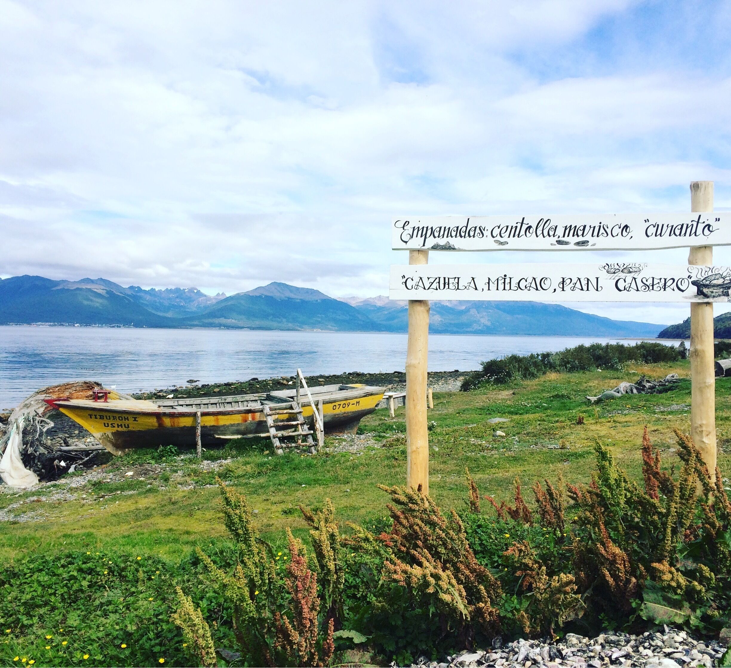 A coastal landscape with a sign listing local delicacies, a boat, and mountains in the background.
