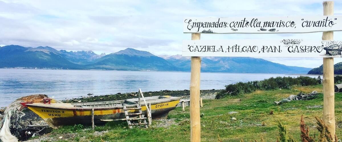 A coastal landscape with a sign listing local delicacies, a boat, and mountains in the background.