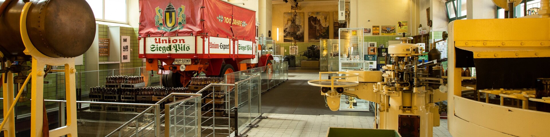 Interior de uma cervejaria, com equipamentos de produção, barris e um grande caminhão vermelho com a inscrição 'Union Siegf. Wils'.