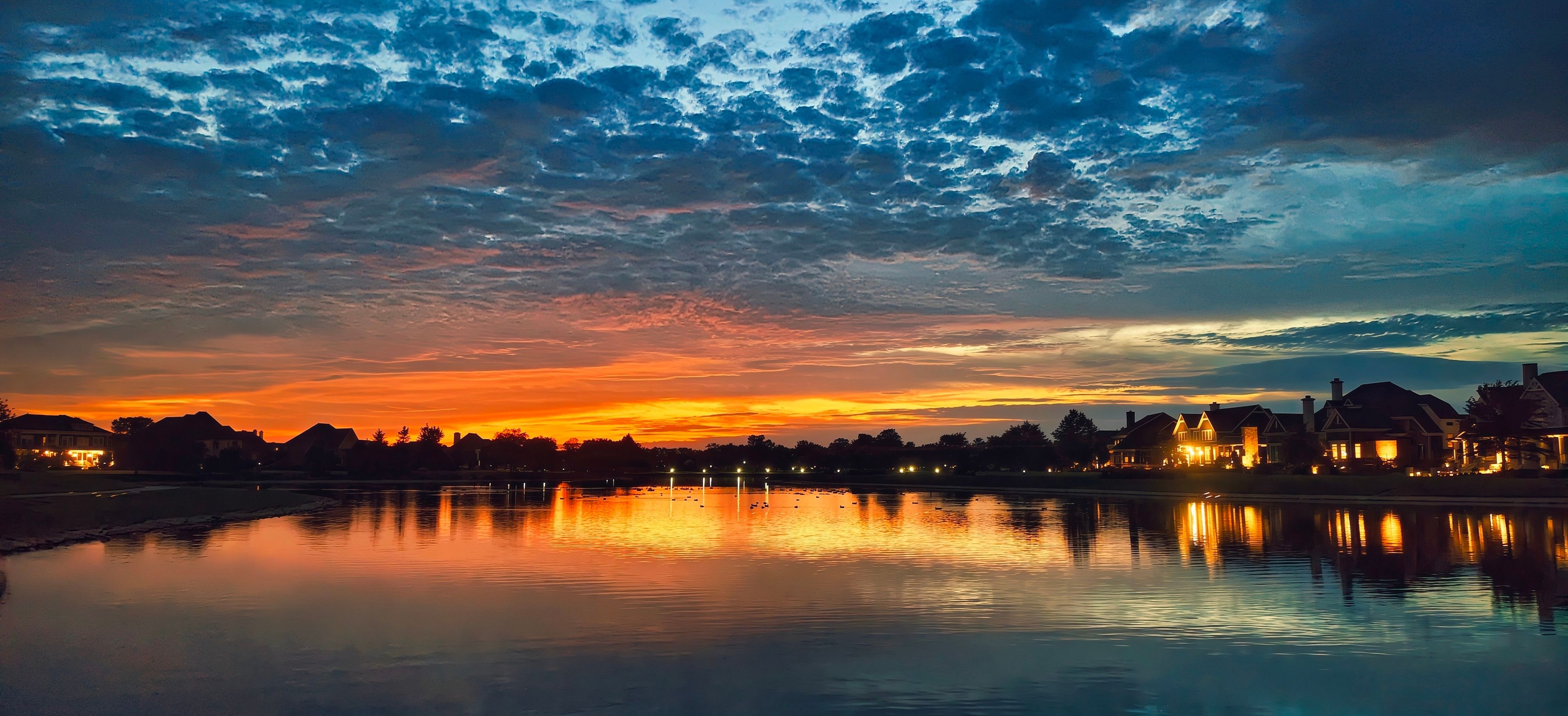 Un atardecer sobre un lago tranquilo, con reflejos de edificios y árboles.