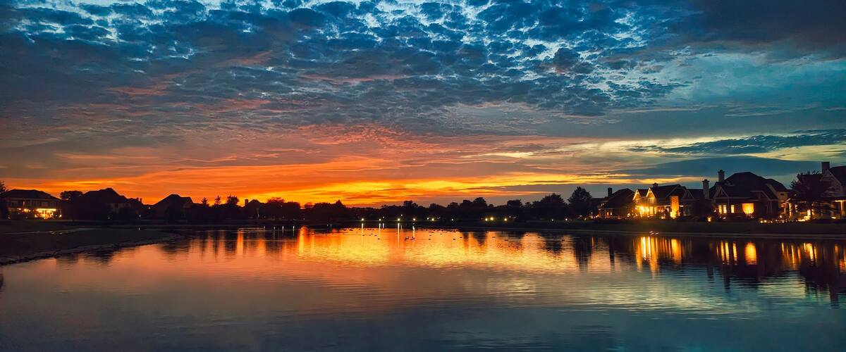 Un coucher de soleil sur un lac paisible, avec les reflets des bâtiments et des arbres qui se mirent dans l’eau.