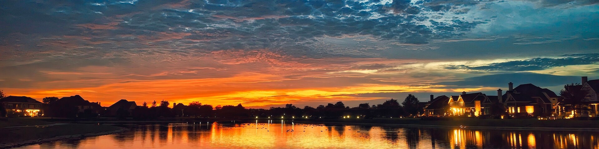 Un atardecer sobre un lago tranquilo, con reflejos de edificios y árboles.