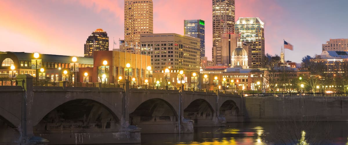 A city skyline at dusk with illuminated buildings, a bridge, and a river reflecting lights.